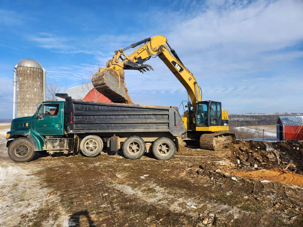 A yellow excavator loads dirt into a large black dump truck on a farm. There are silos and barns in the background, and the ground is partially covered with snow and dirt under a blue sky.