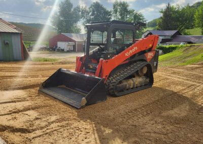 A bright orange Kubota skid steer loader sits on freshly graded dirt near barns and trees on a sunny day.