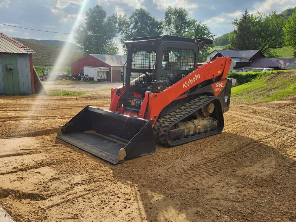 A bright orange Kubota skid steer loader sits on freshly graded dirt near barns and trees on a sunny day.