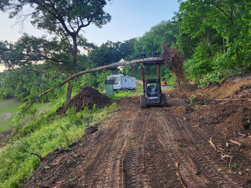 A small skid-steer loader lifts a large uprooted tree on a dirt path surrounded by green trees and brush, with a white trailer visible in the background. The scene appears to be a clearing or construction site.