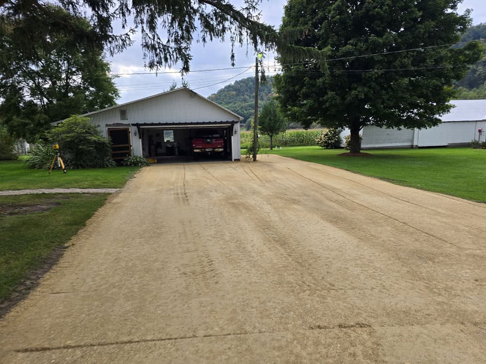 A freshly smoothed gravel driveway leads to a white garage with an open door, where a red vehicle is parked inside. Lush green grass, trees, and a white outbuilding surround the area.