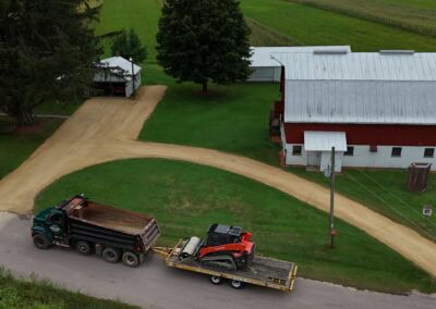 Aerial view of a red and white barn with a gravel driveway on green grass. A truck with a trailer carrying construction equipment is parked on the road near the barn. Trees and fields surround the property.