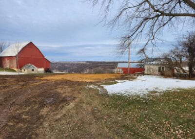 A rural scene with a large red barn, an old stone building, sparse patches of snow on the ground, muddy paths, leafless trees, and rolling hills in the background under a cloudy sky.
