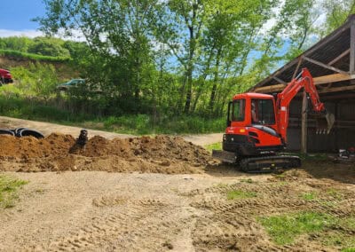 A small red excavator is parked next to a pile of dirt near a partially open shed, with trees, grass, and parked cars in the background on a sunny day.