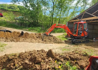 A small red excavator is digging a trench near a partially constructed or open-sided building, with trees and greenery in the background under a blue sky. Dirt piles are scattered around the excavation site.
