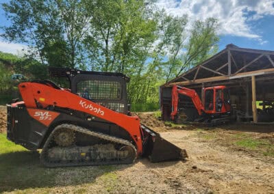 A bright orange Kubota skid steer loader is parked on a dirt path near a metal shed, with another orange excavator partially visible inside the shed. Green trees and blue sky fill the background.