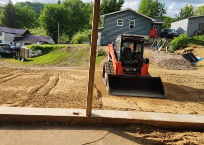 A small orange bulldozer is leveling dirt in a residential yard, viewed from a covered patio. Green trees, houses, and parked cars are in the background under a partly cloudy sky.
