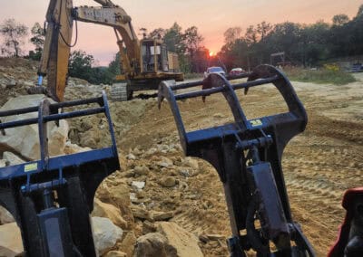 A construction site at sunset with a large excavator moving rocks and dirt. The foreground shows the metal arms of construction equipment, and trees are visible in the background under a colorful sky.