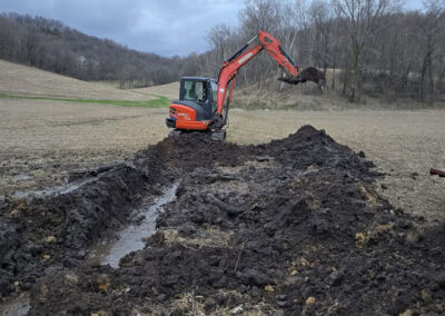A red excavator digs a long trench in a muddy, open field surrounded by bare trees and hills under a cloudy sky.