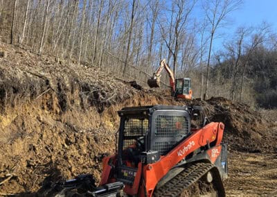 A red Kubota compact track loader with a grapple attachment sits on muddy ground at a construction site, with a small excavator working on a slope in the background and bare trees under a clear blue sky.