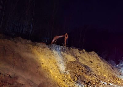 An excavator sits atop a steep dirt embankment at night, illuminated by bright artificial lights, with bare trees visible in the background against the dark sky.