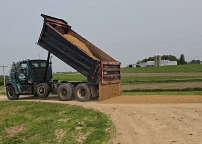 A dump truck unloads a pile of gravel onto a rural dirt road, with farmland, green fields, and a few farm buildings visible in the background under a clear sky.
