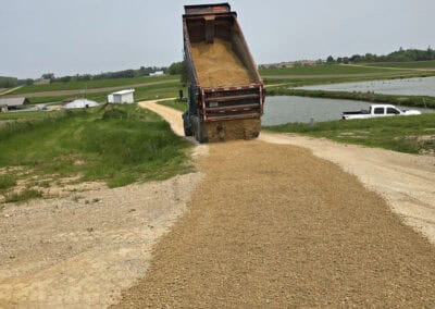 A dump truck spreads a load of gravel onto a rural dirt road next to a pond, with green fields and buildings in the background under a clear sky.