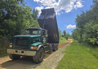 A green dump truck with its bed raised is parked on a dirt road surrounded by trees and grass under a blue sky with scattered clouds.
