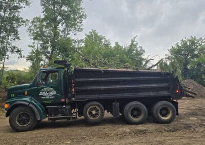 A green and black dump truck carrying branches and debris is parked on a dirt lot, surrounded by trees and piles of soil under a cloudy sky.