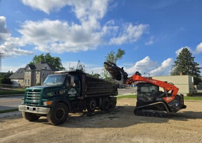 A black dump truck is parked on a gravel lot while an orange Kubota skid steer loader lifts dirt into the truck's bed under a blue sky with scattered clouds. Houses and trees are visible in the background.