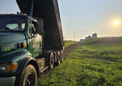 A green dump truck with its bed raised is parked on a grassy field under a clear sky with the sun low on the horizon. Silos and farm buildings are visible in the distance.