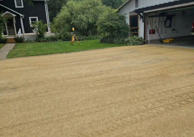 A gravel road runs in front of a black house and a white garage. A yellow tripod survey instrument stands on the green lawn between the buildings. Trees and a cloudy sky are visible in the background.