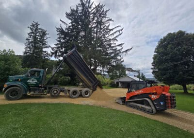 A dump truck unloads gravel onto a driveway while an orange skid steer loader spreads the material. The scene takes place outdoors with grass, trees, and a white building in the background.