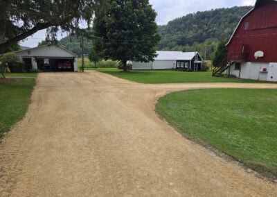 A dirt driveway forks between a red barn with an outdoor basketball hoop and two white farm buildings, surrounded by green grass, trees, and hills in the background.