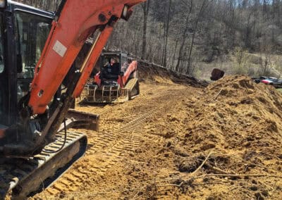 Two orange Kubota excavators are parked on a construction site with sandy soil and tire tracks. Trees without leaves line the edge of the site under a clear blue sky.