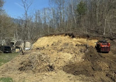 A dirt hillside is being excavated near a wooded area. A red excavator is parked on the right, and a house with a parked car is visible on the left. The sky is clear and blue.