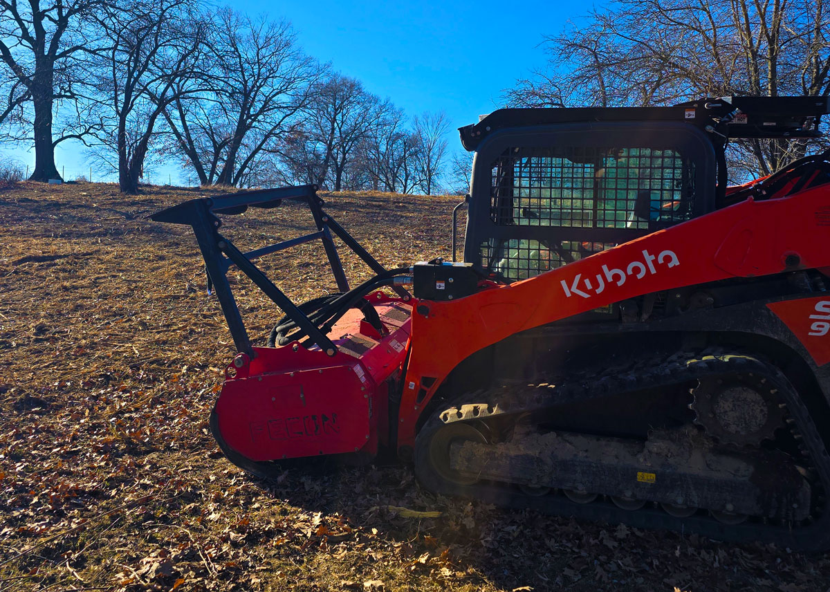 A red Kubota skid steer loader with a front attachment is parked on a grassy, leaf-covered hill under a clear blue sky, with bare trees in the background.