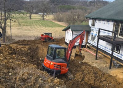 A small excavator digs near a house under construction, with another construction vehicle in the background on a dirt-covered yard surrounded by trees and hills.