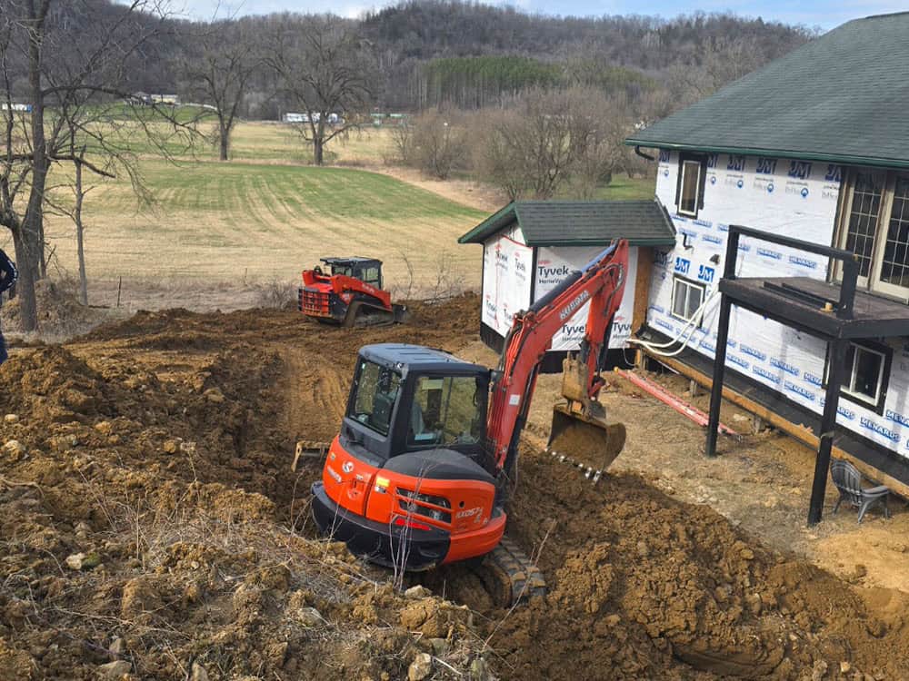 A small excavator digs near a house under construction, with another construction vehicle in the background on a dirt-covered yard surrounded by trees and hills.