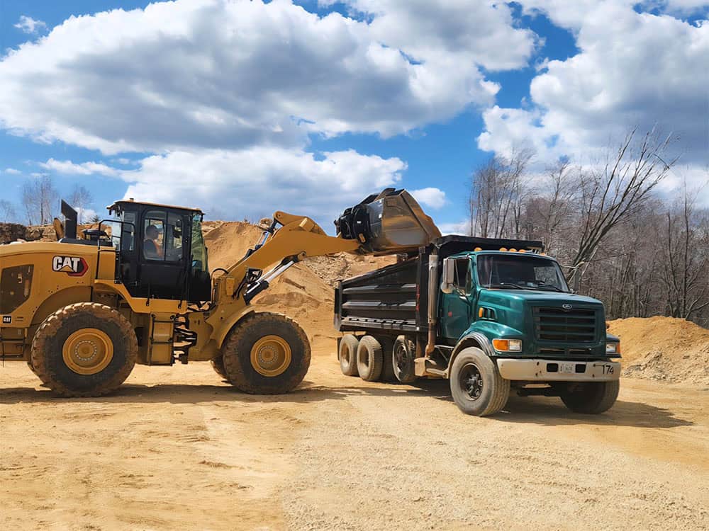 A yellow front loader fills the bed of a green dump truck with sand at a construction site under a partly cloudy sky. Dirt piles and leafless trees are in the background.