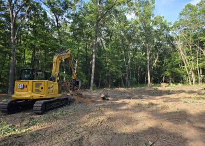 A yellow excavator sits on cleared dirt next to a forested area, with felled trees and scattered branches visible around the edge of the clearing.