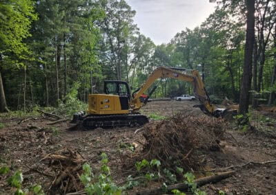 A yellow excavator moves a pile of uprooted branches in a clearing surrounded by trees, with a white pickup truck parked in the background.