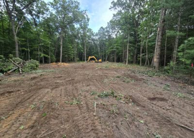A cleared, muddy area in a forest with scattered branches and a yellow excavator in the background, surrounded by tall green trees under a partly cloudy sky.