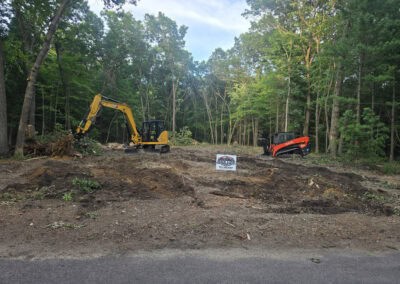 A small excavator and skid steer sit on cleared forest land near a roadside, surrounded by tall trees. A sign in front reads “Anderson Contracting, LLC.” The ground is covered with dirt and uprooted vegetation.