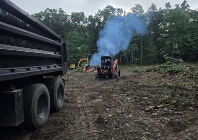 A construction site with heavy machinery, including a dump truck and a loader emitting blue smoke, on cleared dirt ground. Trees line the background under a cloudy sky.
