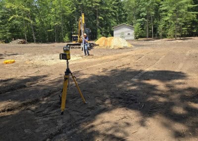 A construction site in a wooded area; a survey tripod is in the foreground, and an excavator with workers and a pile of dirt are near a small gray shed in the background under a blue sky.