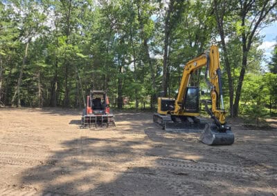 A red skid steer and a yellow excavator sit on cleared dirt ground, surrounded by tall trees and greenery under a partly cloudy sky.