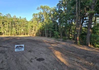 A cleared, dirt lot bordered by dense trees under a blue sky. A real estate sign is posted in the foreground, indicating the land is available for sale. Sunlight shines through the trees onto the ground.