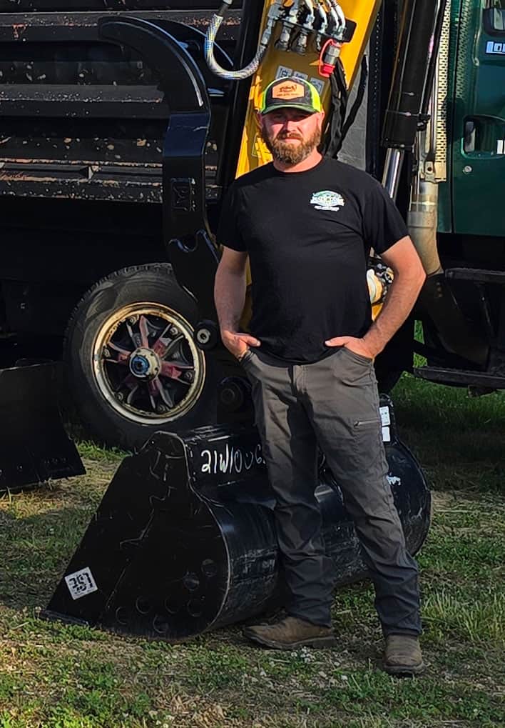 kaleb-mack A bearded man in a black T-shirt, gray pants, and a yellow cap stands outdoors, leaning against construction equipment with a black bucket attachment and a truck wheel visible behind him.