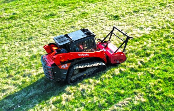 A red Kubota skid steer with a Fecon mulcher attachment sits on a grassy field, casting a shadow on the ground.